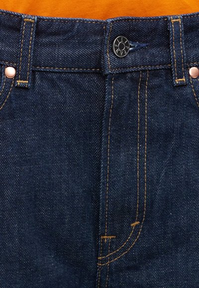 Jean en denim bleu foncé avec des surpiqûres orange, des rivets en cuivre et un bouton en métal noir sur un fond en tissu orange.