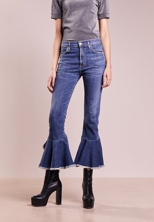 Persoon die blauwe denim jeans draagt met uitlopende en rafelige zomen, gecombineerd met zwarte plateauhakken en een grijs T-shirt met korte mouwen.