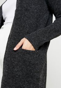 Długi, ciemnoszary kardigan z miękkiego, teksturowanego materiału. Posiada boczne kieszenie i prążkowane mankiety, co zapewnia luźny krój.