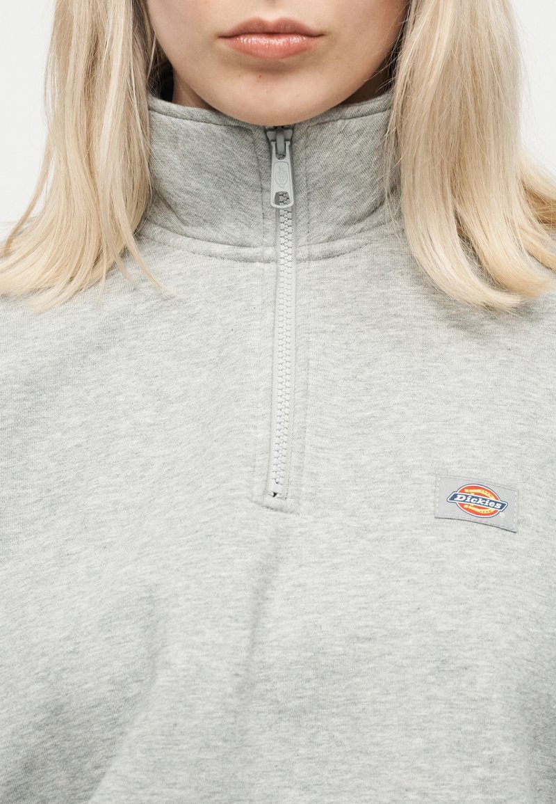 Pullover grigio chiaro con cerniera a quarti e collo alto indossato da una persona con capelli biondi lisci; piccola toppa con logo Dickies sul petto.