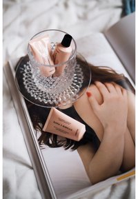 Drei rosa Kosmetiktuben in einem Kristallglas auf einem klaren Teller, der auf einer Zeitschrift mit einem Foto einer Frauenhand liegt.