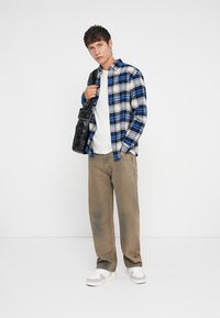 Camisa de flanela xadrez azul e branca sobre uma t-shirt branca, combinada com calças largas bege e ténis brancos, segurando uma mala de ombro preta.