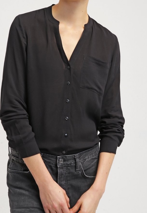 Personne portant une chemise noire à manches longues avec une poche poitrine et un jean gris foncé, les mains légèrement jointes devant.