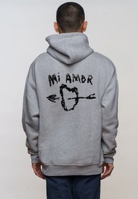 Sudadera gris con un gran gráfico negro en la parte posterior que presenta el texto "Mi Amor" y un corazón con flechas, hecha de un material suave.