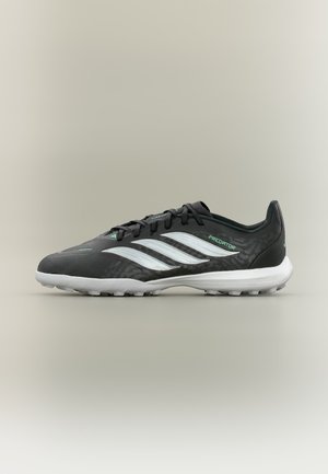 Chaussure de football Adidas Predator noire avec des rayures blanches, des accents verts, un design à lacets et une semelle en caoutchouc blanche sur fond beige.