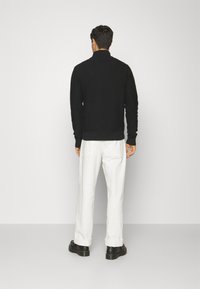 Pulover negru tricotat cu guler înalt, finisaj texturat și tiv elastic, asortat cu pantaloni albi și pantofi negri.