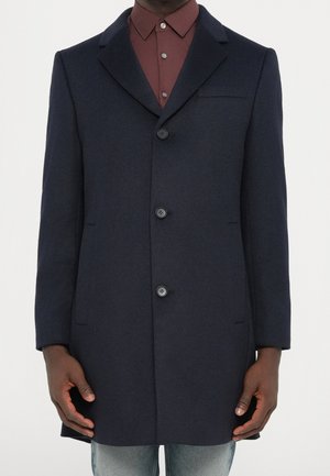 Homme portant un manteau bleu marine foncé à simple boutonnage avec trois boutons sur une chemise bordeaux boutonnée et un jean bleu clair.