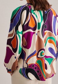 Blus med långa ärmar i lätt tyg, prydd med ett flerfärgat abstrakt mönster med virvlar i lila, grön, orange och bruna nyanser.