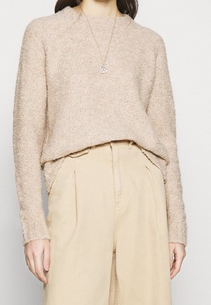 Frau trägt einen beigen, strukturierten Pullover, der in hoch taillierte cremefarbene Hosen gesteckt ist, accessorisiert mit einer runden Anhänger-Halskette.