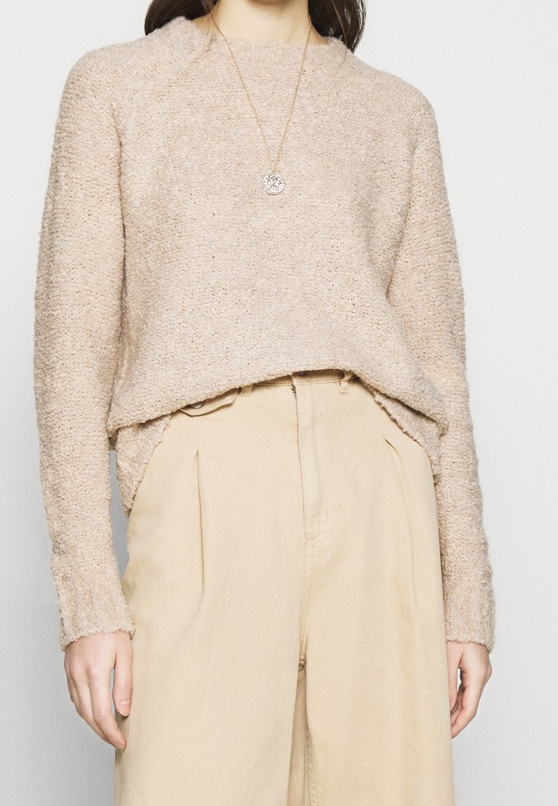 Femme portant un pull texturé beige rentré dans un pantalon crème taille haute, accessoirisée d'un collier pendentif rond.