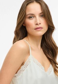 Camisole ivoire avec bordure en dentelle au niveau du décolleté et des bretelles. Texture lisse, tissu léger, avec un motif délicat en dentelle.