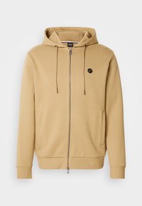 Hnedá mikina na zips vyrobená z mäkkej bavlnenej zmesi. Má kapucňu so sťahovacou šnúrkou, bočné vrecká a malý logotyp na hrudi.