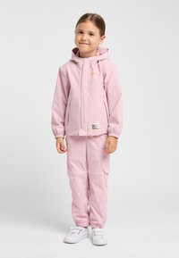 Lichtroze waterproof jack met capuchon en voorzakken, gecombineerd met lichtroze cargobroek met elastische taille. Gemaakt met witte sneakers.