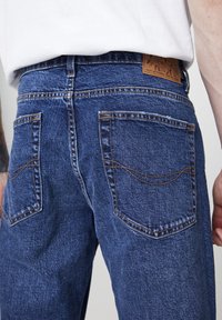 Pantalones vaqueros de mezclilla azul con diseño de pierna recta, costuras visibles, dos bolsillos traseros y una etiqueta de cuero marrón claro con la marca en la cintura.