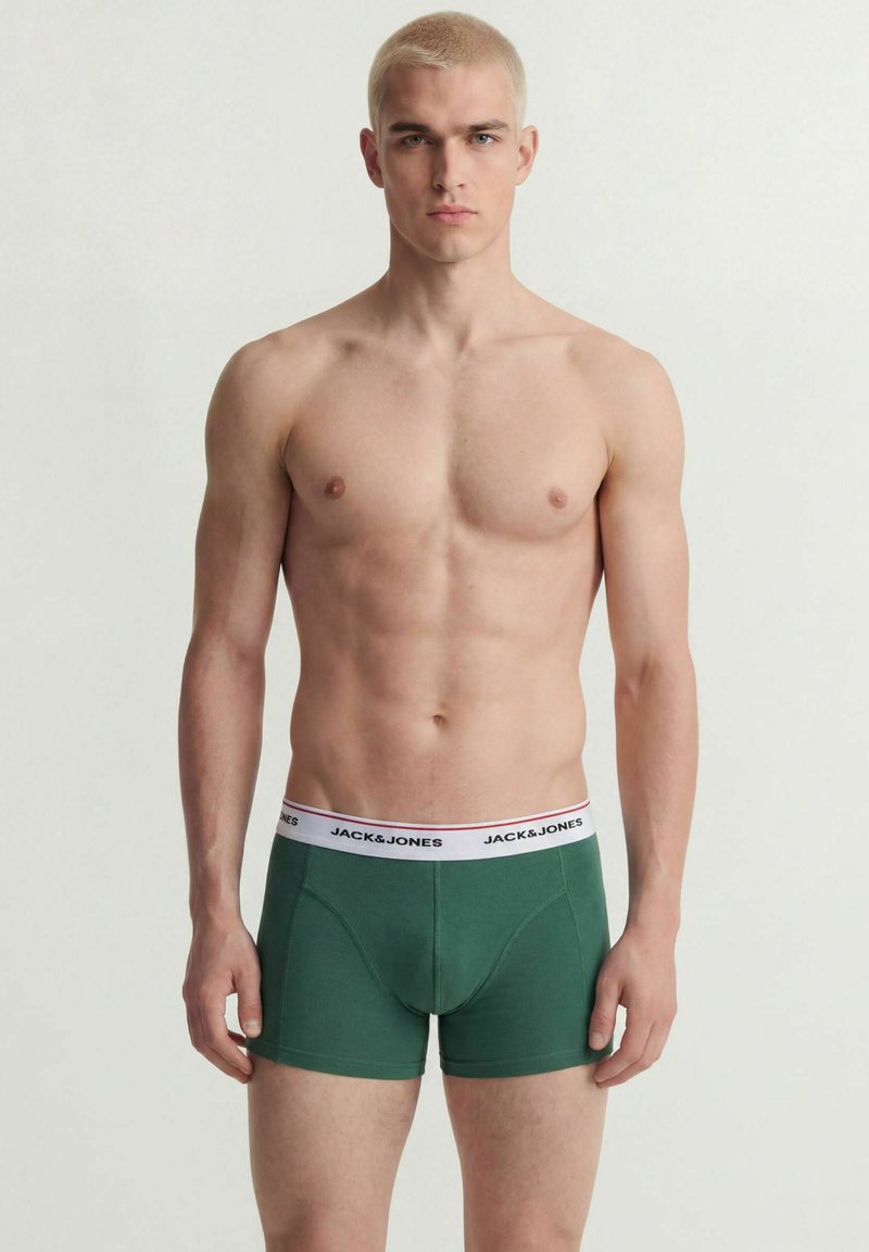 Jeune homme aux cheveux blonds courts, debout face à l'avant, portant un boxer vert avec une ceinture blanche marquée sur un fond uni.