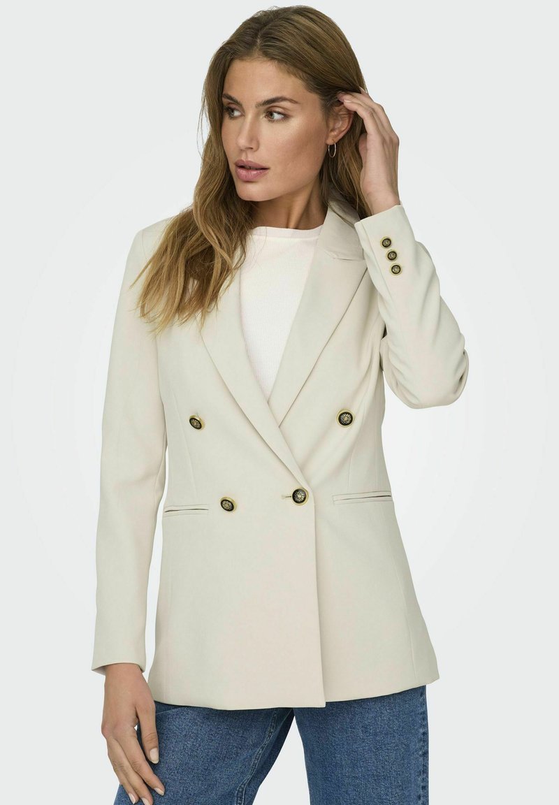 Blazer doppiopetto beige con dettagli in bottoni dorati, spalle strutturate e tasche laterali; indossato sopra una maglietta bianca e jeans blu.