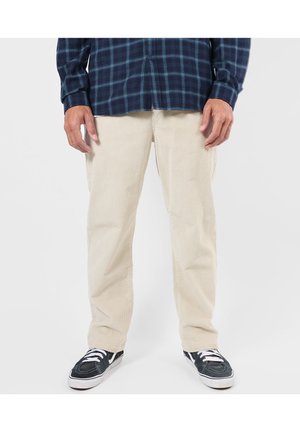 Homme portant un pantalon en velours côtelé beige, des baskets noires et blanches, et une chemise à carreaux bleu foncé, debout devant un fond uni.