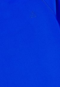 Material textil de un albastru strălucitor, care prezintă o textură netedă. Deține un logo subtil într-o nuanță mai deschisă de albastru, amplasat în partea stângă sus.