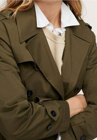 Olivengrøn trenchcoat med store klipper, dobbelbrystede knapper og optrukne ærmer over en cremefarvet sweater og hvid skjortekrave.