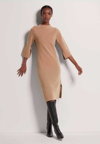 Beige Strickkleid mit hohem Kragen, ausladenden Ärmeln und einem seitlichen Schlitz, kombiniert mit schwarzen kniehohen Stiefeln mit Absatz. Gesamte glatte Textur.