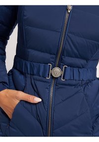 Mano parcialmente en el bolsillo de una chaqueta acolchada azul con cremallera central y un cinturón de hebilla de la marca Guess alrededor de la cintura.