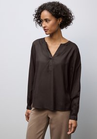 Blouse à manches longues, marron foncé, en tissu doux avec un col en V et des poignets froncés, associée à un pantalon beige. Design simple et épuré.