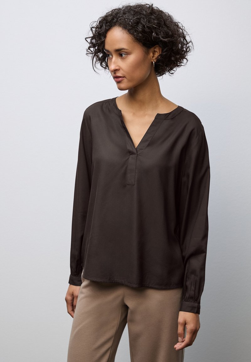 Blouse à manches longues, marron foncé, en tissu doux avec un col en V et des poignets froncés, associée à un pantalon beige. Design simple et épuré.