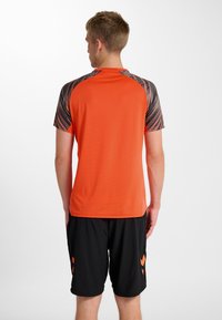 Orange kortärmad sporttröja med en texturerad rygg och ett svart mönster på axlarna, bärs tillsammans med svarta shorts.