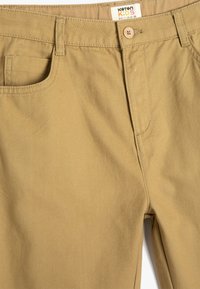 Pantalones de niño color caqui hechos de algodón, con una textura suave, diseño tradicional de cinco bolsillos, cierre de botón y etiqueta de la marca en la cinturilla.