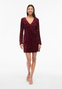 Burgundská šatka s flitrami, dlhými rukávmi, výstrihom do V a priliehavým strihom. Látka má textúrovaný, lesklý vzor a krátky lem.