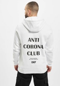 Weißer Hoodie mit schwarzem Text auf dem Rücken, der "ANTI CORONA CLUB" und zusätzliche Nachrichten liest. Verfügt über eine verstellbare Kapuze und gerippte Bündchen.