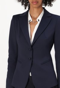 Marineblå skreddersydd blazer med to knapper, klassiske revers og klaffelommer; båret over en hvit bluse med dekorative detaljer.
