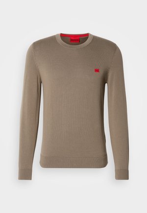 Suéter de punto beige con mangas largas, cuello redondo, puños y dobladillo acanalados, que presenta un pequeño parche de logo rojo en el pecho.