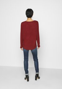 Haut en maille bordeaux à manches longues, coupe décontractée, ourlet arrondi, associé à un jean bleu ajusté et des bottines noires à talons, style minimaliste.
