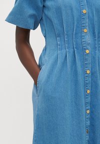 Denim skjortklänning i ljusblått med korta ärmar, sidofickor, samlad midja och guldknappar. Slät textur och avslappnad design.