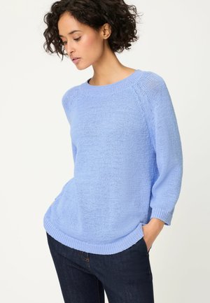Frau mit lockigem Haar, die einen hellblauen Strickpullover und dunkle Jeans trägt, Hände in den Taschen, blickt nach unten zur Seite.