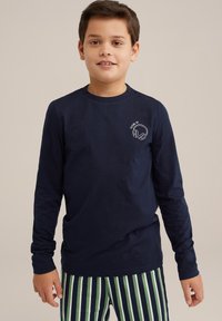 Mornarska majica s dolgimi rokavi iz bombaža, z majhnim belim grafičnim prikazom slušalk na prsih. V kombinaciji z zelenimi in belimi prugastimi hlačami.