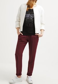 Veste bomber matelassée blanche sur une chemise noire avec un design graphique argenté, associée à un pantalon tapered bordeaux et des baskets blanches.