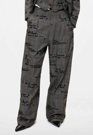 Pantaloni a quadri a vita alta e gamba larga con dettagli in scrittura nera manoscritta, indossati con blazer abbinato e scarpe nere a punta.