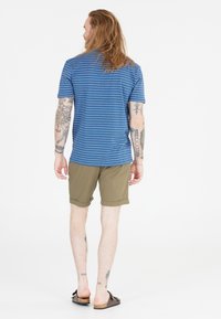 Blå randig kortärmad skjorta, beige upprullade shorts och bruna slip-on skor. Synliga tatueringar på armar och ben. Neutral bakgrund.