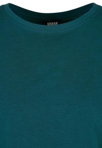 Turquoise katoenen T-shirt met ronde hals, zichtbaar stiksel langs de kraag en een zwart etiket met de tekst "URBAN" aan de binnenzijde van de halslijn.