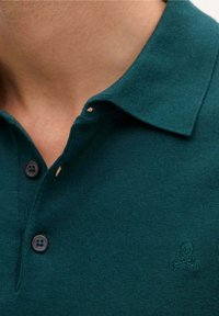 Polo skjorte i mørkegrøn strikket stof, med en knapkrave og to mørke knapper, prydet med et lille broderet dødningehoved og korslagte knogler logo.