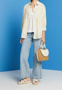 Cremefarbenes, marmoriertes Hemd mit Knöpfen, weißes V-Ausschnitt-Oberteil, hellblaue Jeans mit weitem Bein, beige Riemchensandalen und eine gewebte Handtasche mit weißen Akzenten.