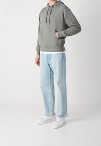 Felpa grigia con cappuccio e tasca frontale, logo bianco, abbinata a jeans a gamba larga blu chiaro e sneakers bianche. Design semplice e casual.