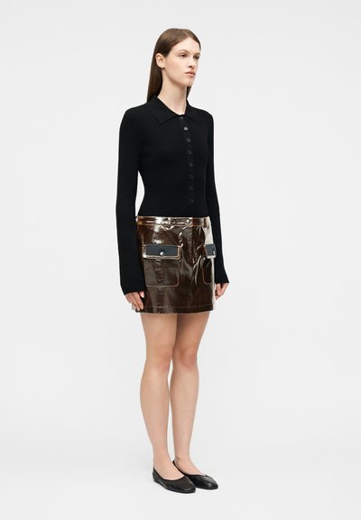 Femme debout portant un haut noir à manches longues boutonné, une mini-jupe marron brillante avec des poches, et des ballerines noires sur un fond uni.