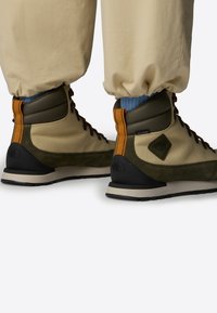 Beżowe i ciemnozielone buty trekkingowe z czarnymi podeszwami, wyposażone w sznurowadła, pomarańczowy akcent i teksturowaną powierzchnię. Wykonane z trwałego, wodoodpornego materiału.