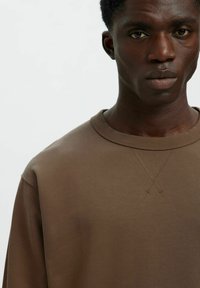 Brūns sporta krekls ar apaļu kakla izgriezumu, smalku krusteniskā dūriena detaļu un mīkstu tekstūru. Audums izskatās gluds un nedaudz pieguļošs.