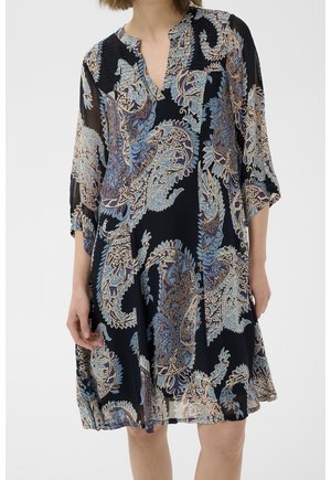 Femme portant une robe ample, longueur genou, avec des motifs cachemire bleus et beiges sur fond noir et manches trois-quarts.