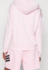 Svetle růžová mikina s kapucí, vyrobená z jemného materiálu a s pružnými manžetami, kombinovaná s odpovídajícími šortkami, které mají tři černé pruhy na boku.