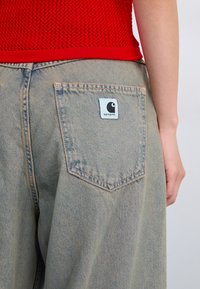 Light blue denim jeans feature a back pocket with a square light blue tag, showcasing the Carhartt logo. The material has a soft, textured finish.